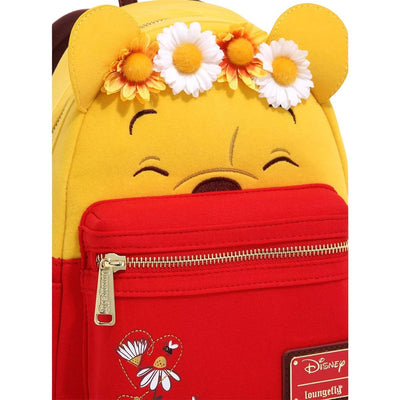 The front view of a yellow mini backpack with a red front pocket. The backpack has a smiling Winnie the Pooh face with a floral crown made of daisies and sunflowers. The red pocket has embroidered flowers and a Disney Loungefly logo.