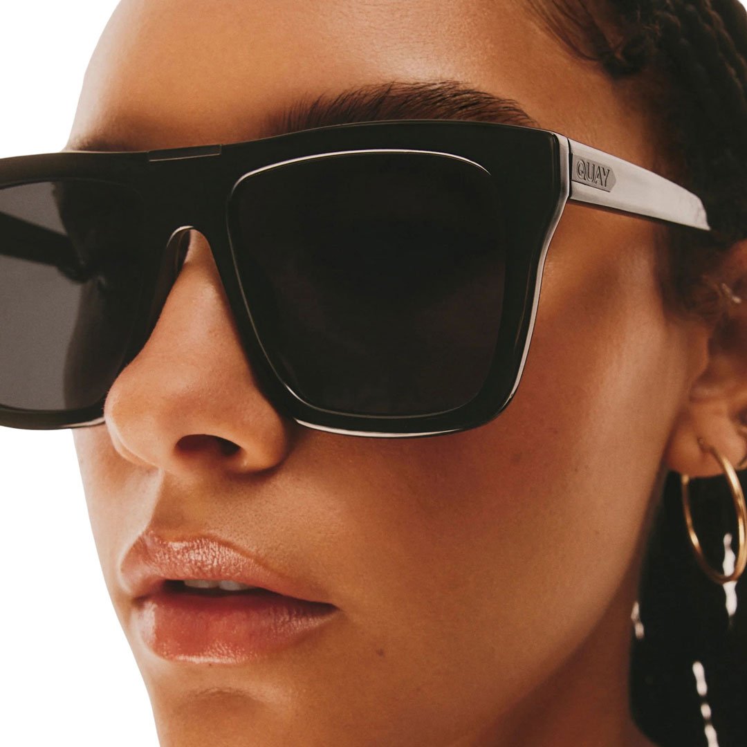 A full-body shot of a woman wearing Quay Australia Women's Name Drop Oversized Square Sunglasses with black frames and black polarized lenses, standing against a white background. She is dressed in a stylish outfit, including a white t-shirt and a black leather corset belt over a black skirt.