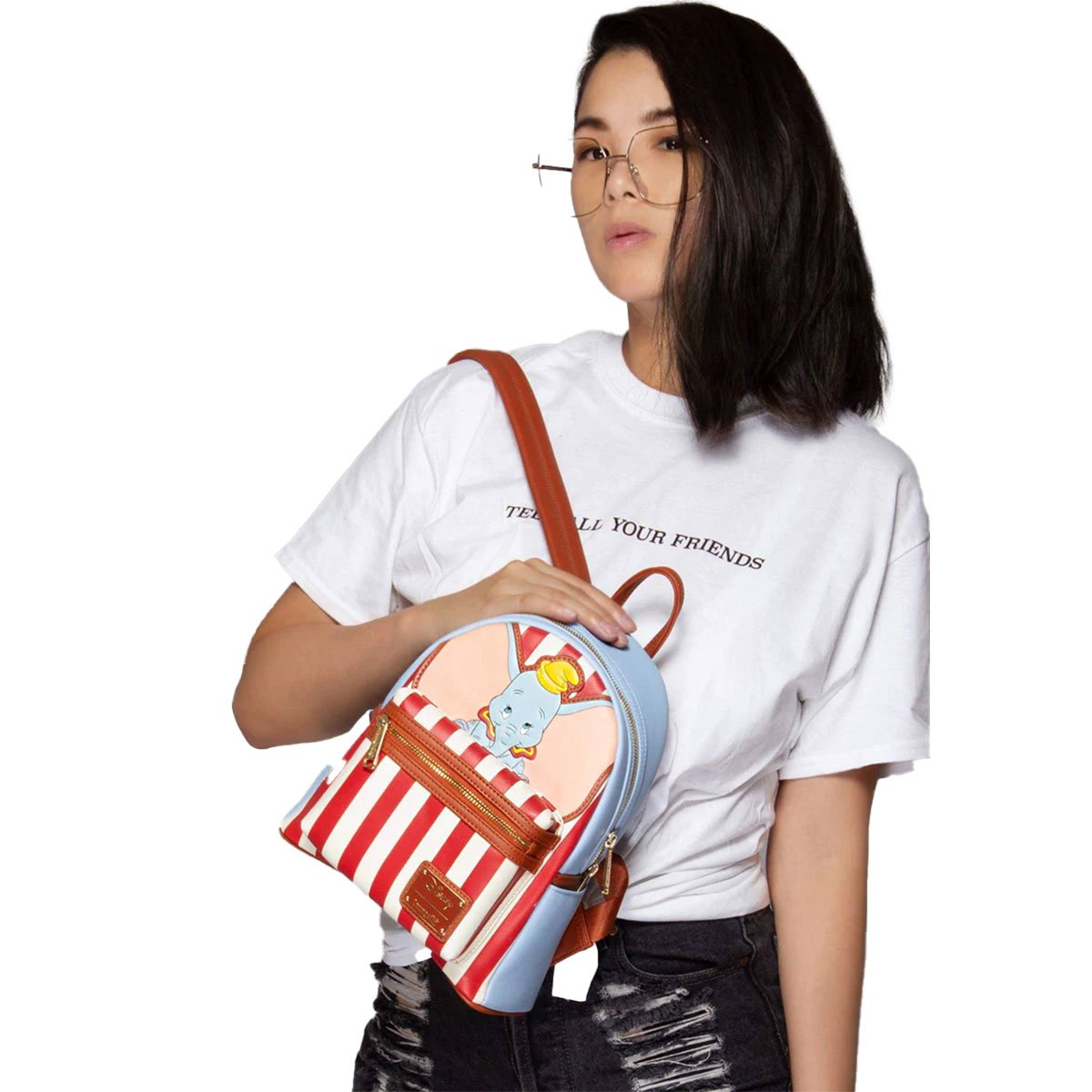 A model wearing the Loungefly Disney Dumbo mini backpack, showing how it fits and looks when worn. The backpack has a light blue exterior, red and white circus stripes, and an image of Dumbo on the front.